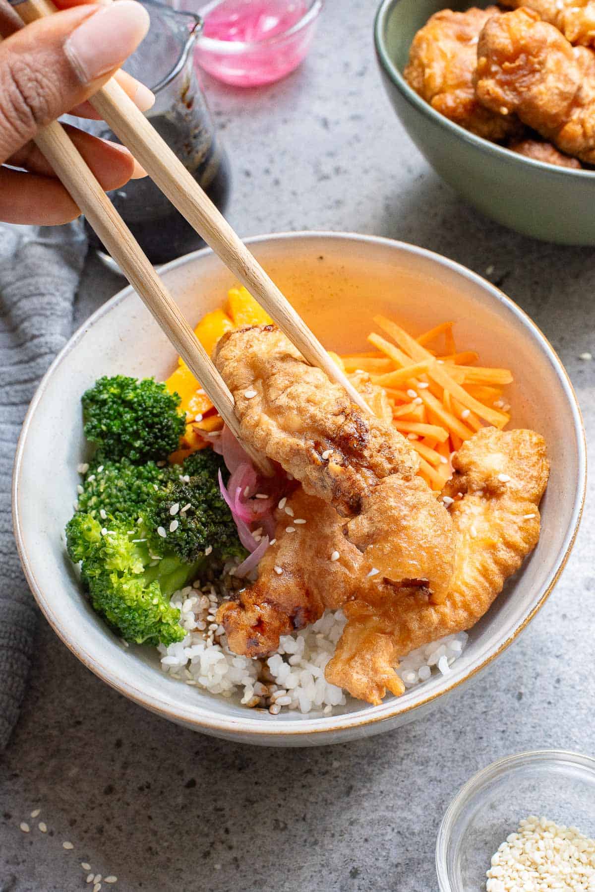 A hand uses chopsticks to hold fried chicken over a bowl filled with rice, broccoli, sliced mango, and carrots, garnished with pickled onions and sesame seeds.