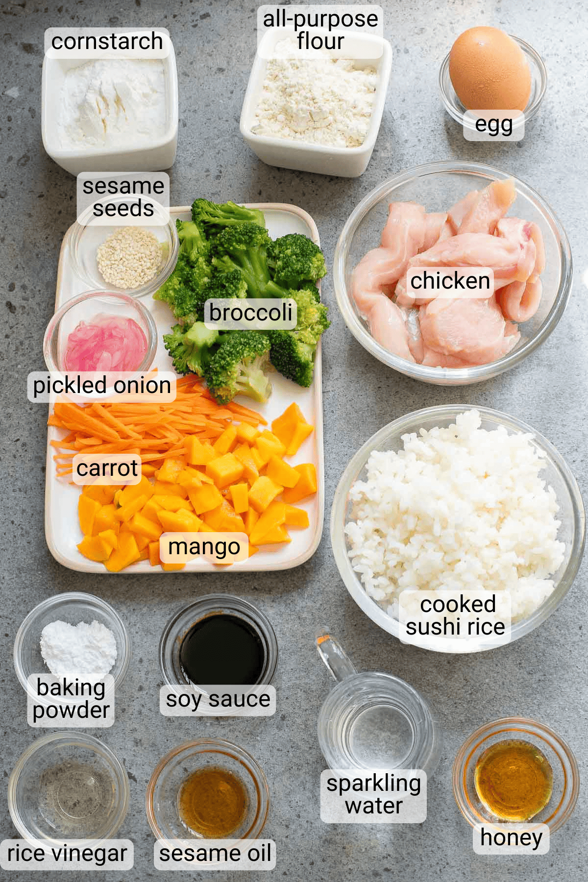 Labeled ingredients on a gray surface include chicken, cooked sushi rice, broccoli, mango, carrot, pickled onion, egg, and various sauces and seasonings.