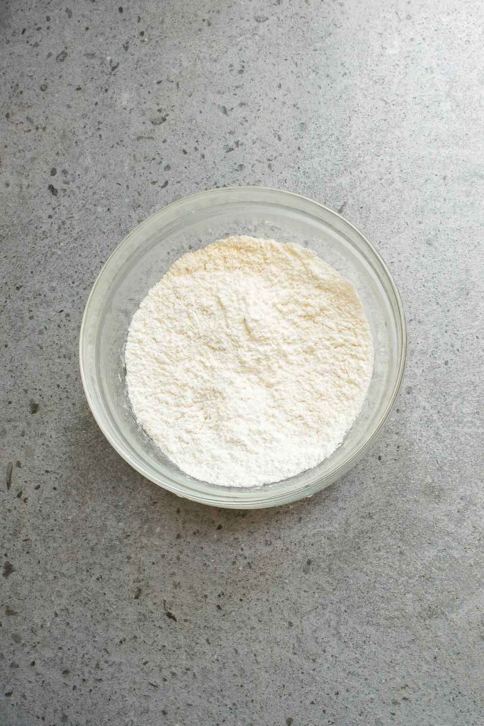 A glass bowl filled with a mound of white flour sits on a gray speckled countertop.