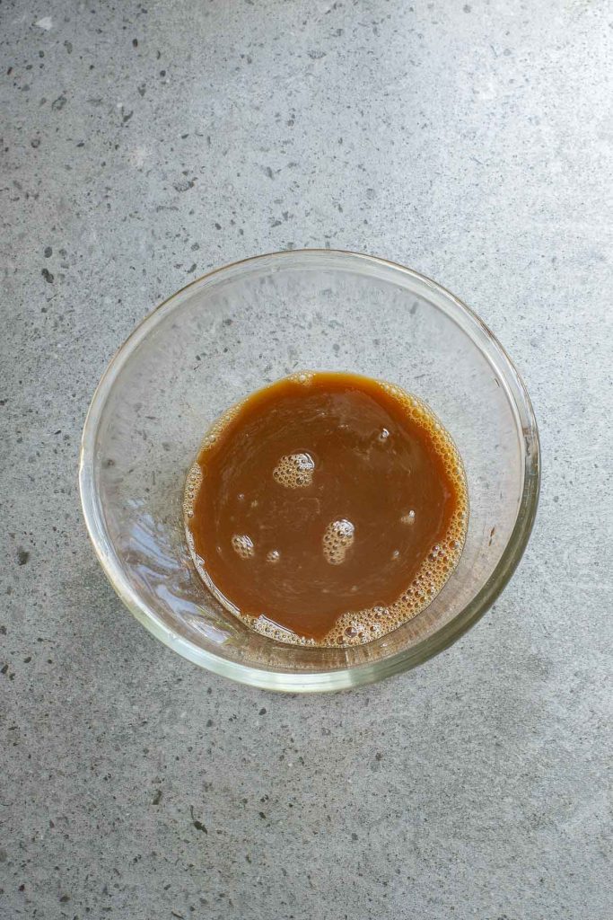 A clear glass bowl containing a brown liquid with bubbles sits on a speckled gray surface.