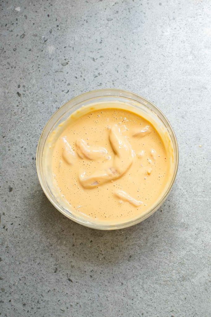 A clear bowl filled with a creamy, yellow batter containing a few elongated pieces, placed on a speckled gray surface.