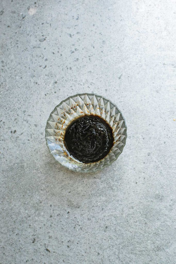 A small glass dish containing a thick, dark sauce sits on a gray, speckled surface.