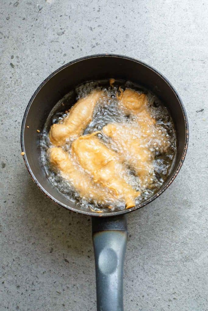 Pieces of battered fish frying in hot oil in a black saucepan.