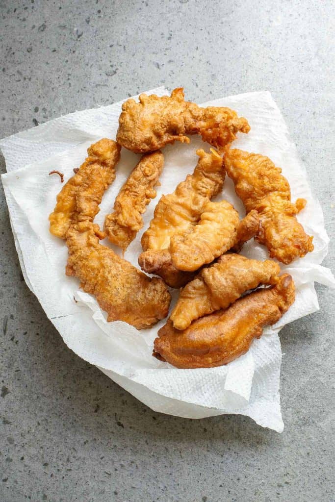 Six pieces of golden-brown fried battered fish on a white paper towel, set on a gray speckled surface.