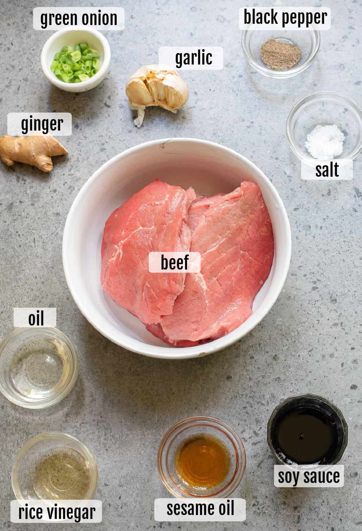 A bowl of raw beef is surrounded by small dishes of green onion, garlic, ginger, black pepper, salt, oil, rice vinegar, sesame oil, and soy sauce, all labeled.