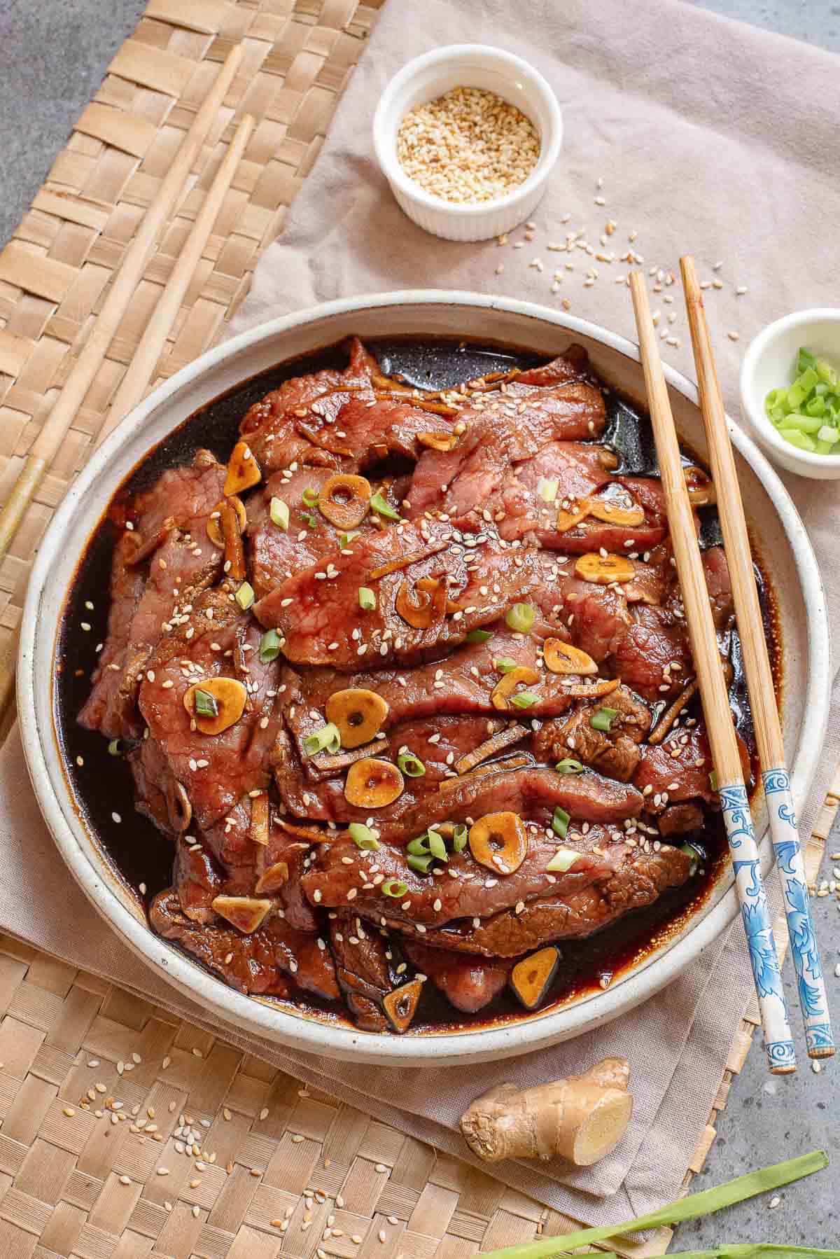 A plate of marinated beef slices garnished with sesame seeds, sliced garlic, and green onions, served with chopsticks. Small bowls of sesame seeds and green onions are nearby.