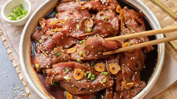 A plate of marinated grilled beef topped with sliced garlic, sesame seeds, and chopped green onions, with chopsticks picking up a piece.