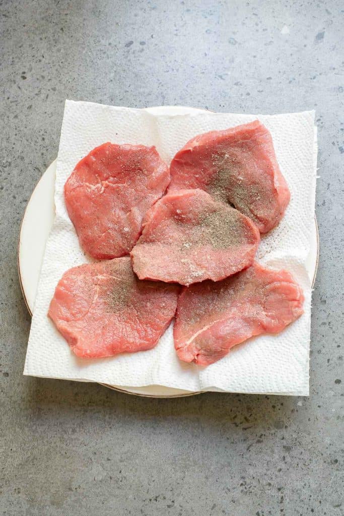 Five raw, thin slices of meat topped with black pepper are arranged on a plate lined with white paper towels on a gray countertop.