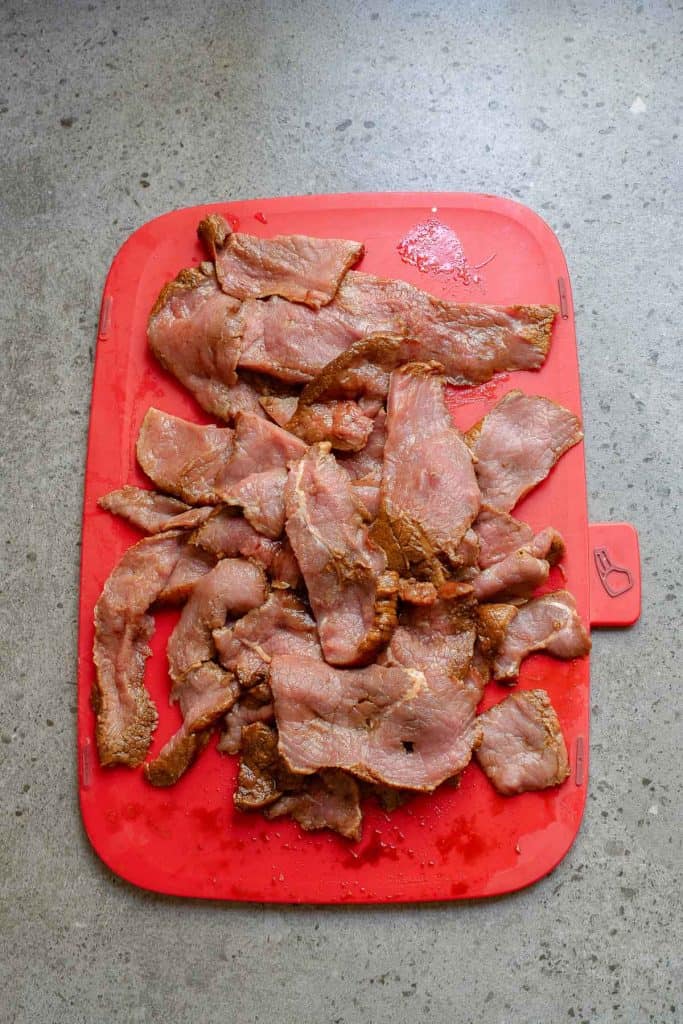 Slices of raw marinated beef are spread out on a red cutting board placed on a gray countertop.