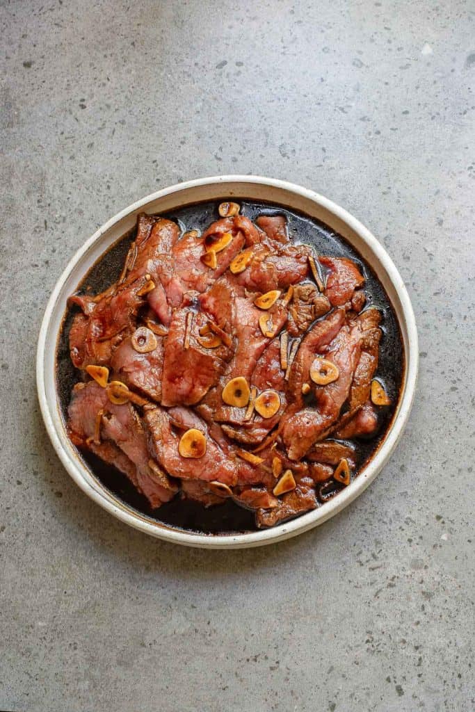 A plate of marinated beef slices topped with sliced garlic, served on a stone-textured surface.