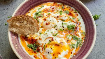 A bowl of poached eggs topped with spiced melted butter, herbs, and black pepper, served with a slice of whole-grain bread.