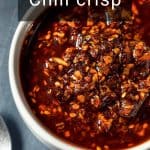 A white bowl filled with chili crisp sauce containing oil, chili flakes, and seeds, placed on a dark surface next to a spoon.