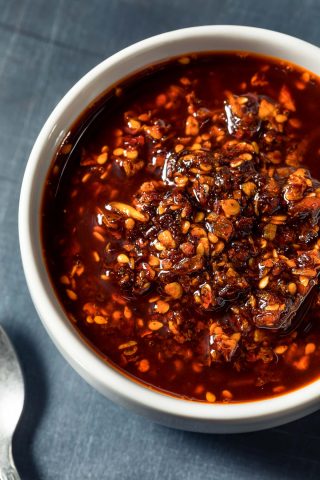 A white bowl filled with chili oil containing visible chili flakes, sesame seeds, and pieces of garlic on a dark surface.