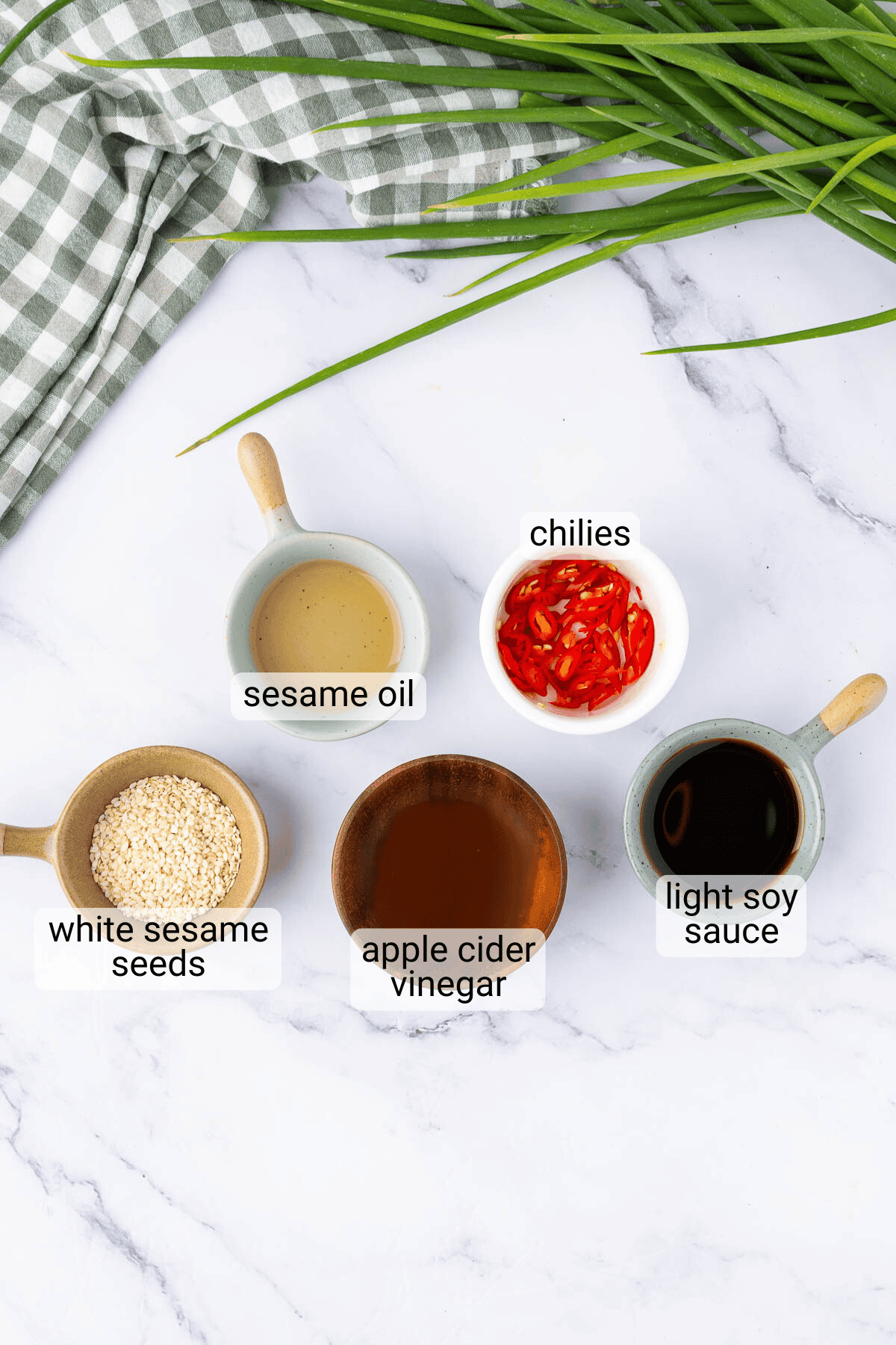 Five labeled bowls containing sesame oil, sliced chilies, white sesame seeds, apple cider vinegar, and light soy sauce on a marble surface with a checkered cloth and chives.