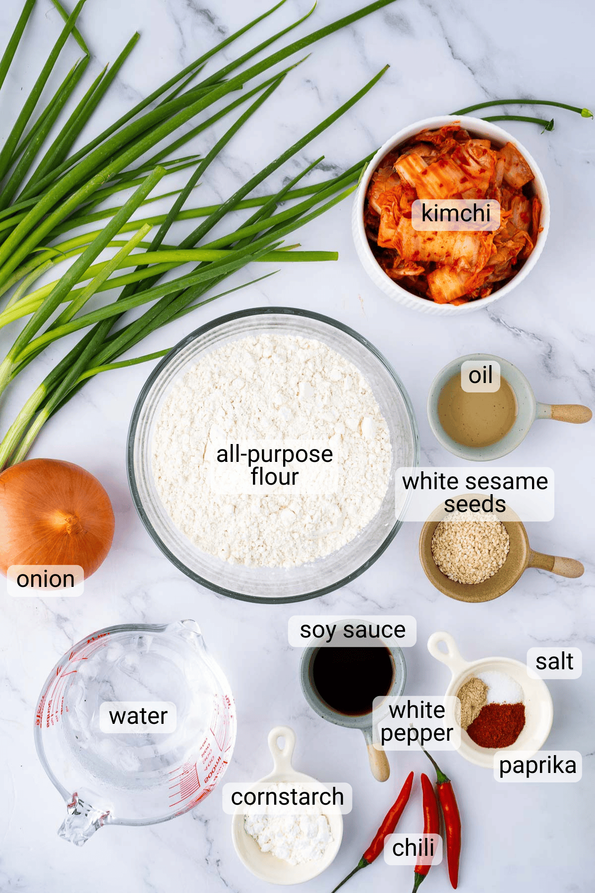 A top-down view of ingredients for kimchi pancakes, including flour, kimchi, green onions, onion, oil, sesame seeds, soy sauce, cornstarch, water, salt, paprika, white pepper, and chili.