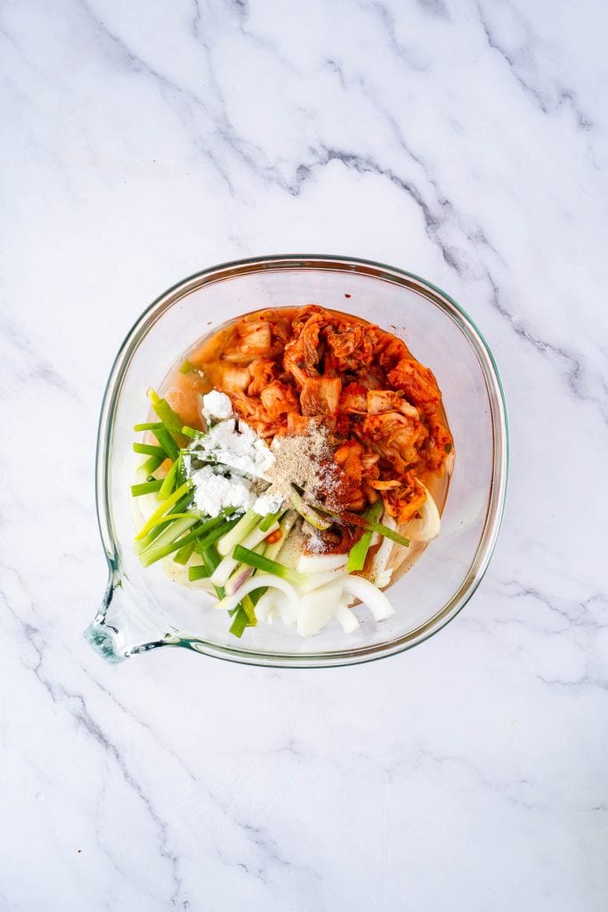 A glass mixing bowl containing kimchi, sliced onions, green onions, white powder (possibly flour or starch), and seasonings on a marble surface.