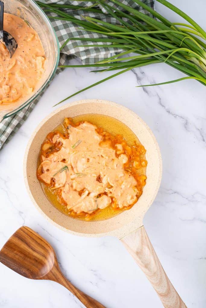 A frying pan with orange batter being cooked on a marble countertop, next to fresh green onions, a wooden spatula, and a bowl containing more batter.