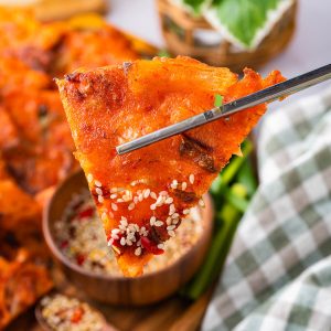 A triangular slice of kimchi pancake is held by metal chopsticks above a bowl of dipping sauce, with a checkered cloth and green garnish in the background.