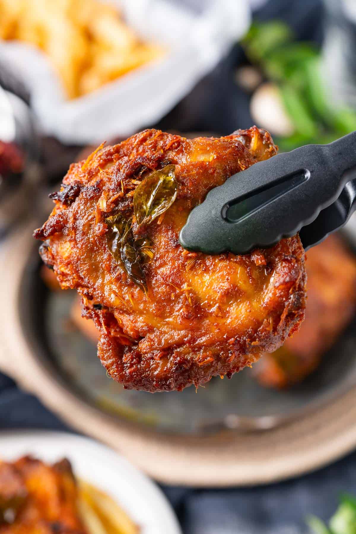 A pair of black tongs holding a piece of crispy, golden-brown fried chicken garnished with herbs, with more chicken pieces visible in the background.