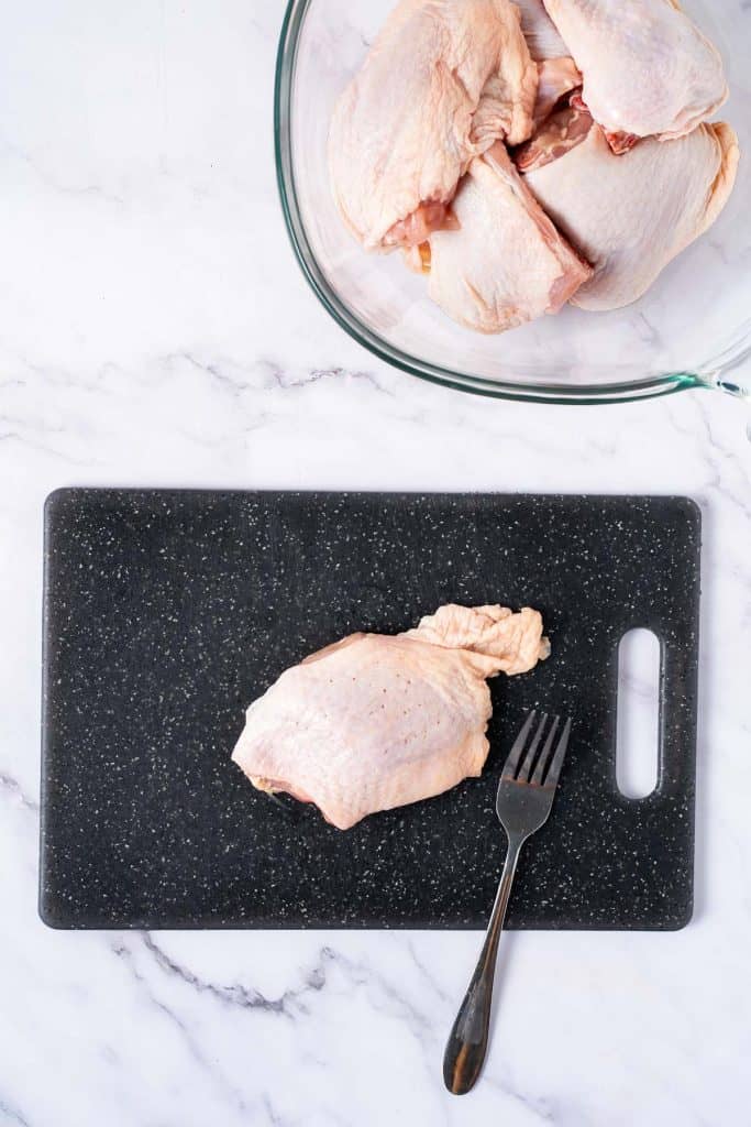 Raw chicken pieces in a glass bowl and one piece placed on a black cutting board with a fork on a marble countertop.