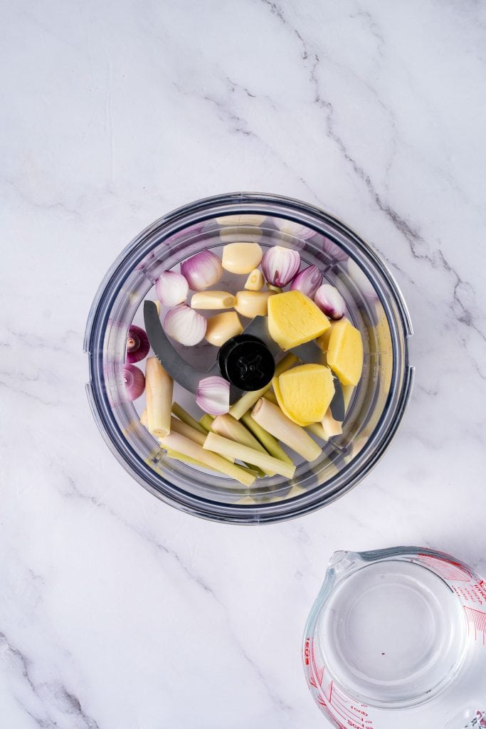 A food processor bowl contains peeled garlic, shallots, lemongrass stalks, and ginger slices; a measuring cup with water is placed nearby on a marble surface.