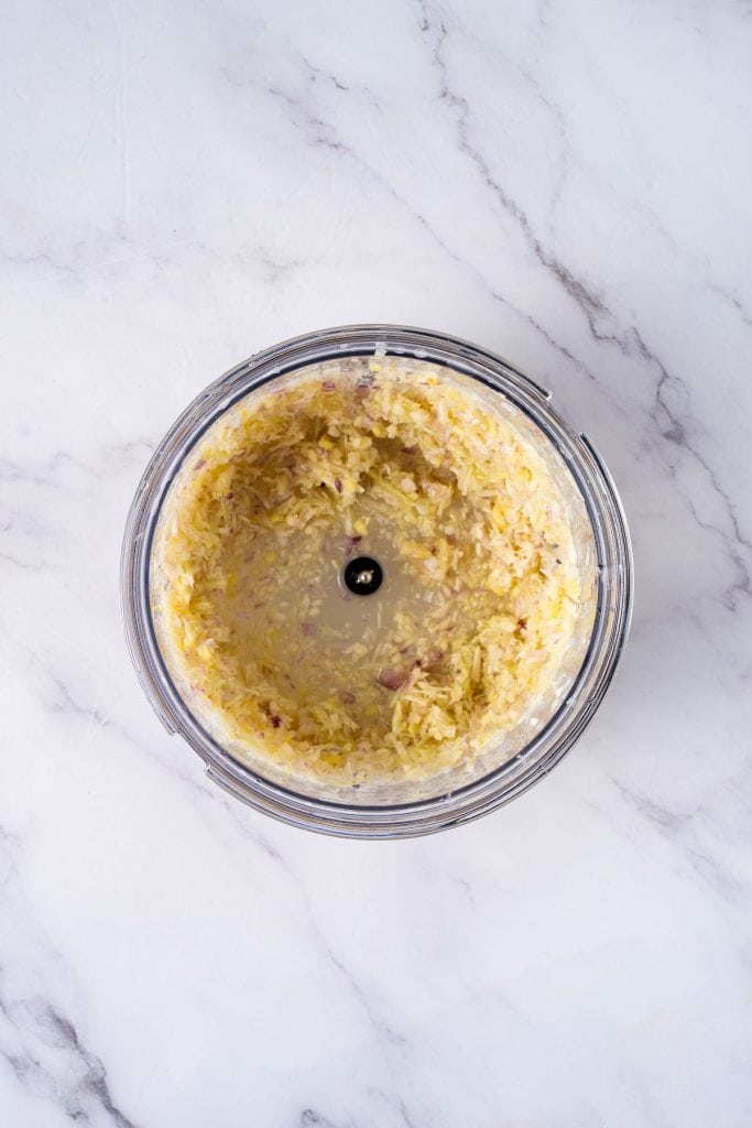 A glass bowl with finely chopped or pureed onions, viewed from above, placed on a white marble surface.