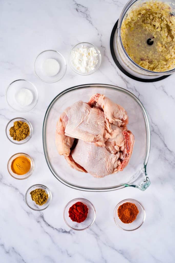 Raw chicken pieces in a glass bowl, surrounded by small bowls of spices, cornstarch, sugar, and a food processor with chopped onions, on a marble countertop.