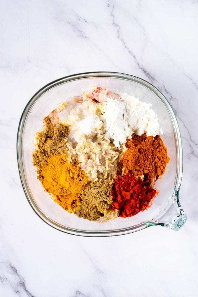 Glass mixing bowl containing ground meat, chopped onions, and various spices including turmeric, paprika, ground coriander, and salt on a white marble surface.