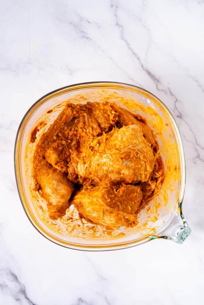 Raw chicken pieces coated in a yellow-orange marinade, placed in a glass mixing bowl on a white marble surface.