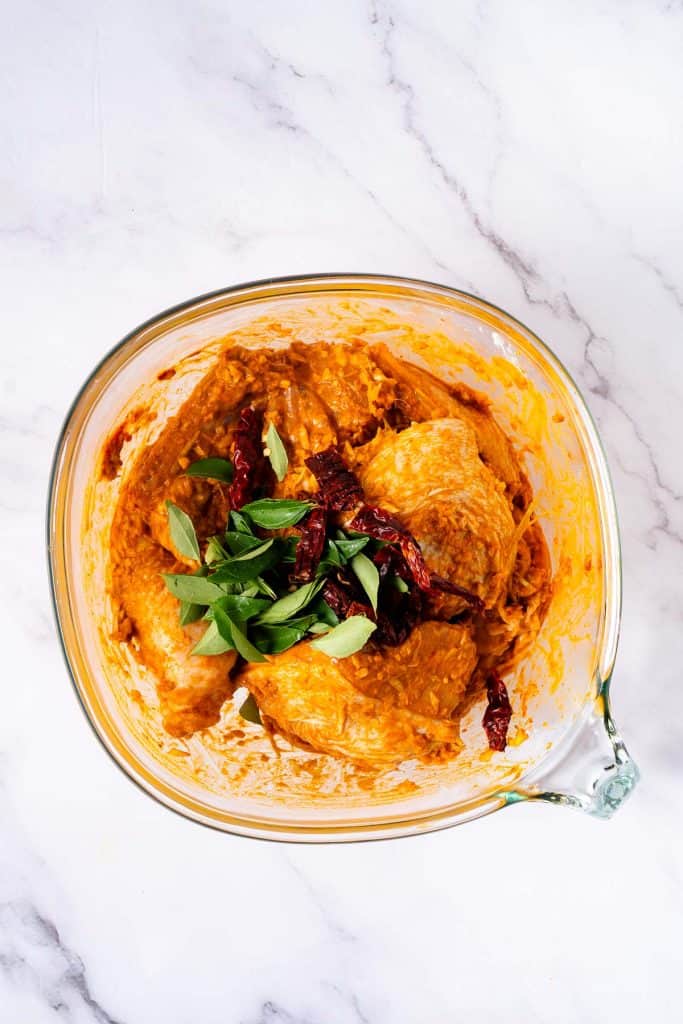 A glass bowl containing chicken pieces coated in an orange marinade, garnished with fresh green herbs and dried red chilies, on a white marble surface.