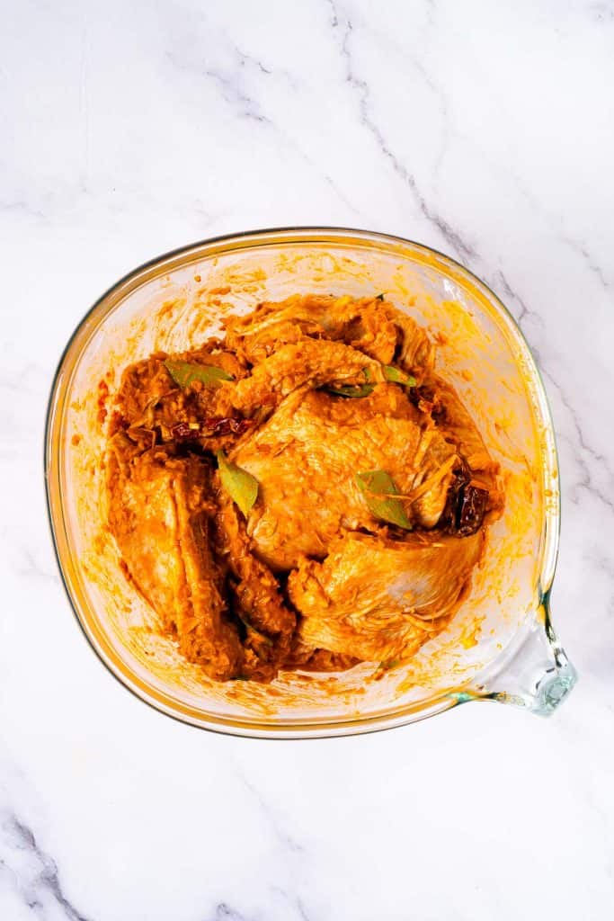 Raw chicken pieces coated in orange marinade with spices and bay leaves in a glass mixing bowl on a white marble surface.