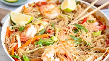 A bowl of stir-fried rice noodles with shrimp, sliced vegetables, green onions, and lime wedges, with chopsticks resting on top.