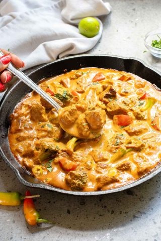 A pan filled with creamy curry containing chunks of meat and vegetables, with a hand holding a serving spoon over the dish.