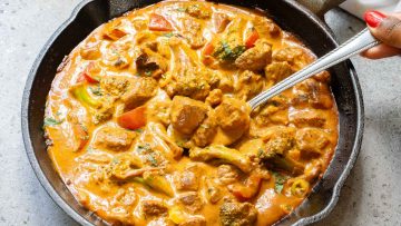 A cast iron skillet filled with creamy curry featuring chunks of meat and vegetables, with a spoon partially submerged in the dish.