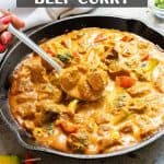 A skillet filled with creamy Thai beef curry, chunks of beef, and vegetables in a rich orange sauce; a spoon scoops some curry.
