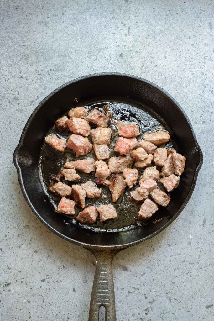 Cubed pieces of beef are being seared in a black cast iron skillet on a gray countertop.