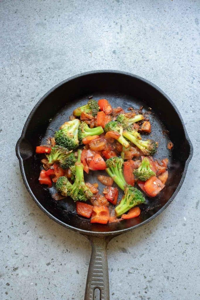 Broccoli and red bell peppers sautéing in a black cast iron skillet on a gray countertop.