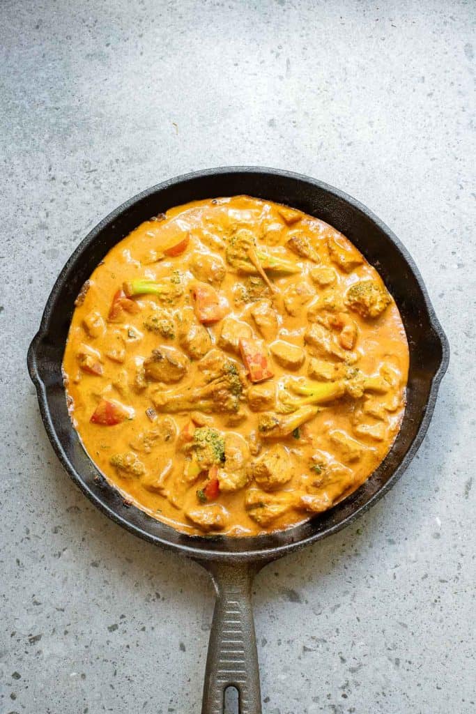 A cast iron skillet filled with a creamy orange curry containing vegetables and chunks of protein, placed on a gray countertop.