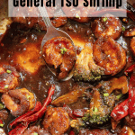 Shrimp cooked in a dark, glossy General Tso sauce with broccoli florets and dried red chilies, served in a pan with a spoon.