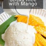 A plate of sticky rice with mango sits on a banana leaf, topped with coconut sauce being poured and garnished with sesame seeds.