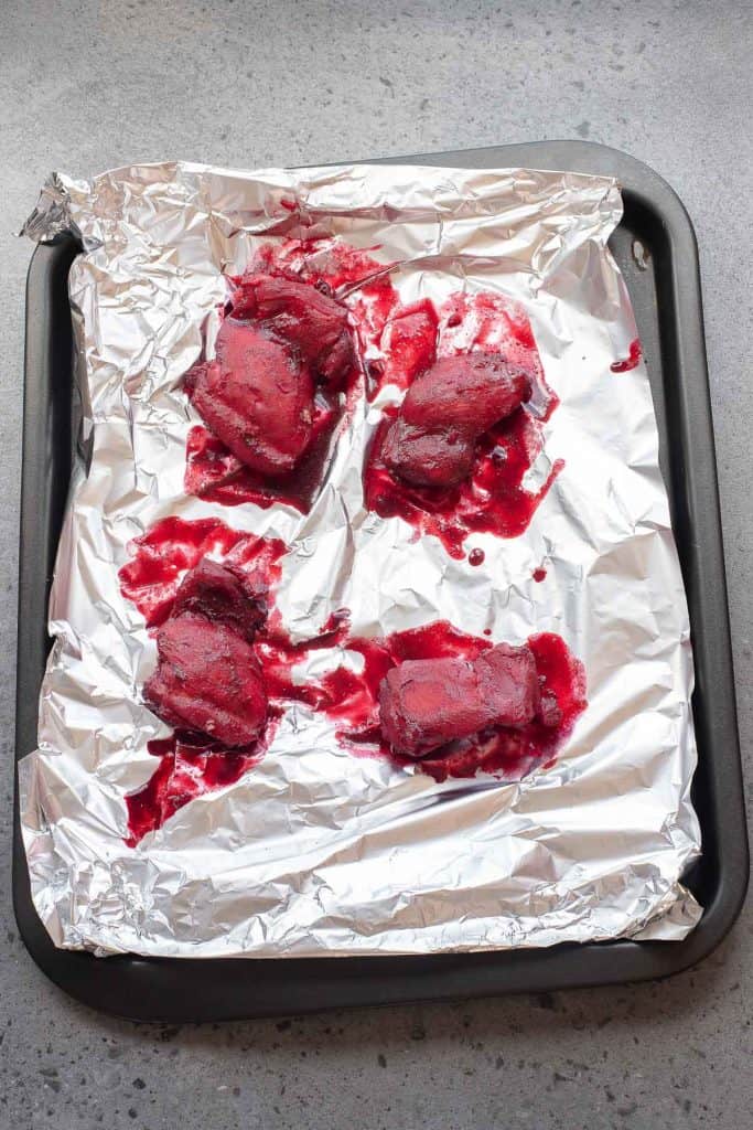 Four pieces of raw, dark red meat are placed on a foil-lined baking tray, with red juices pooling around each piece.