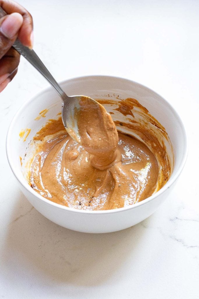 A hand holding a spoon is mixing a thick, light brown batter in a white bowl on a white surface.
