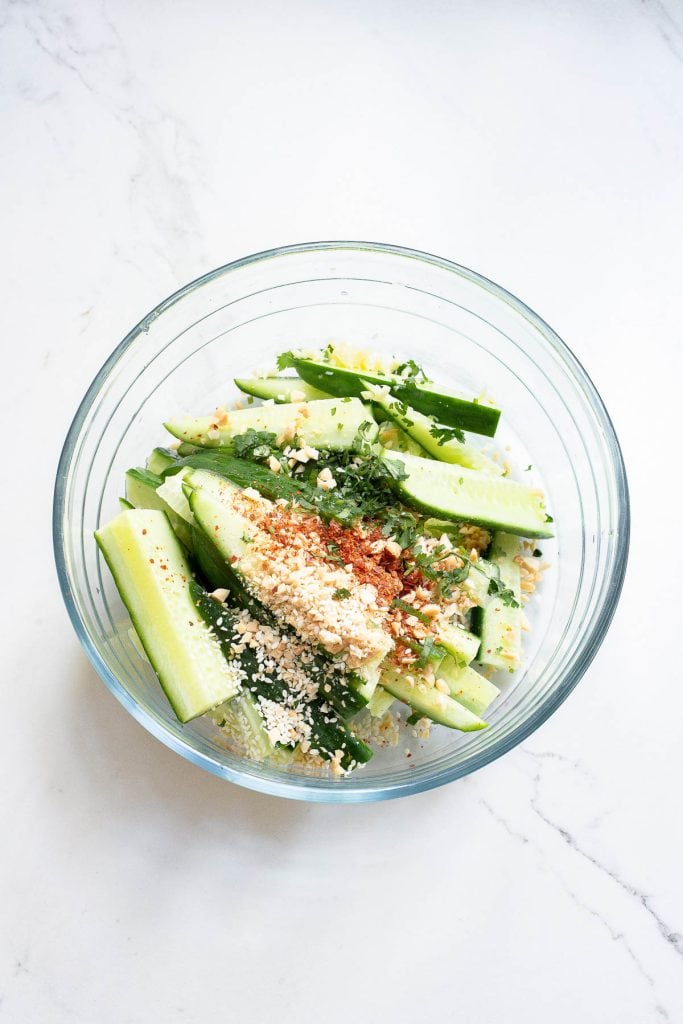 A glass bowl contains sliced cucumbers topped with seasoning, chopped herbs, crushed nuts, and red pepper flakes on a white marble surface.