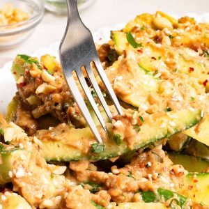 Close-up of a fork pressing into cucumber slices topped with a creamy peanut sauce, chopped peanuts, sesame seeds, and herbs on a white plate.