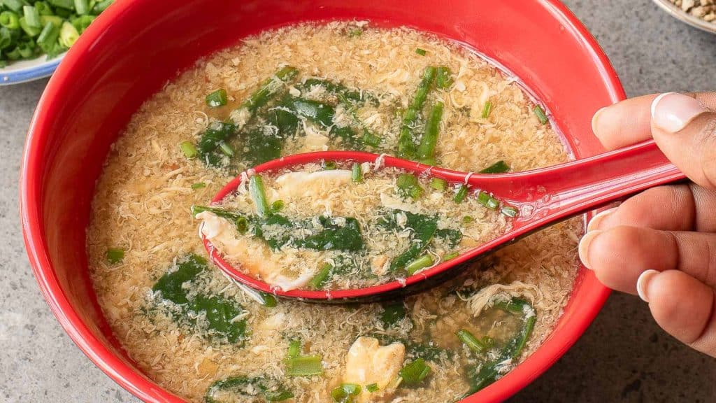 A hand holds a red spoon with egg drop soup, garnished with chopped green onions and leafy greens, above a matching red bowl.