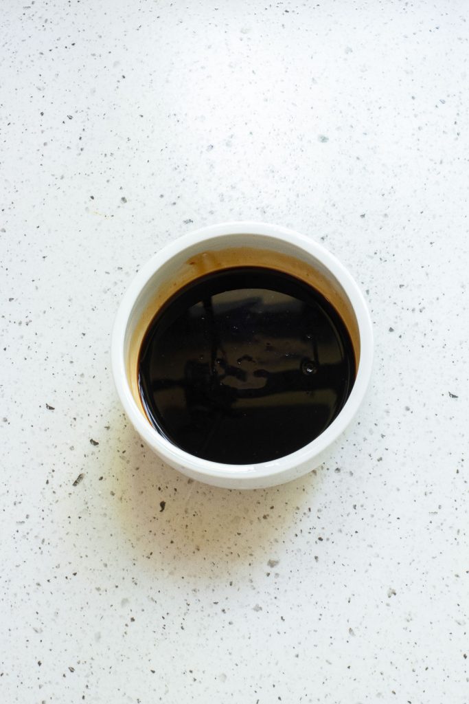 A small white bowl filled with dark soy sauce sits on a speckled countertop.