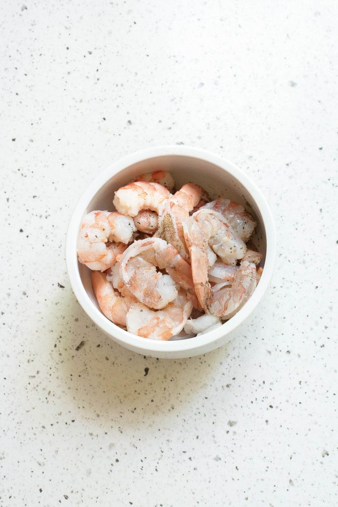 A white bowl filled with raw, peeled, and deveined shrimp sits on a speckled white countertop.
