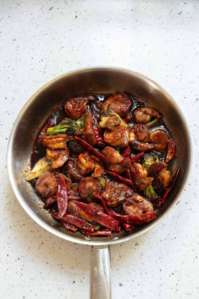 A stainless steel pan on a light countertop contains cooked shrimp, broccoli, dried red chilies, and a dark sauce.