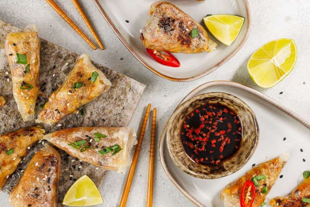 Crispy spring rolls and rice paper dumplings on plates with lime wedges, dipping sauce, sliced red chili, and chopsticks on a light surface.
