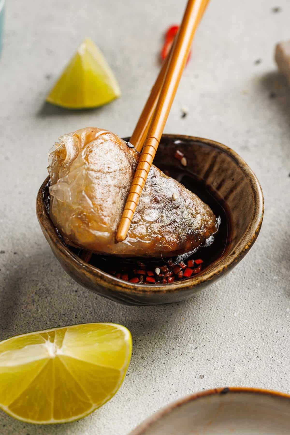 Chopsticks holding a rice paper dumpling over a small bowl of soy sauce with red chili, with lime wedges and rice paper dumplings on a gray surface.
